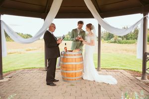 A wedding ceremony takes place under a white-draped gazebo with a wine barrel altar on brick pavers.