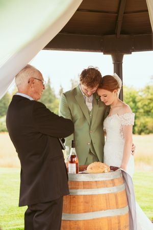 A wedding ceremony takes place under a gazebo with an officiant and couple gathered around a wooden wine barrel.