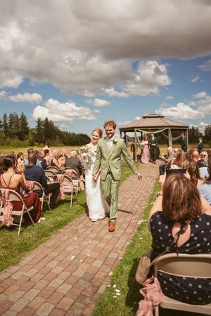 Happy couple walks down a brick aisle between wedding guests under dramatic cloudy skies with a gazebo in the background.