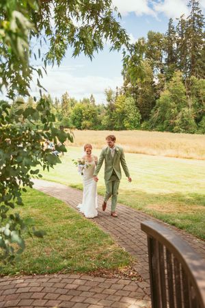 A couple walks along a curved brick pathway surrounded by green trees and manicured lawn on their wedding day.