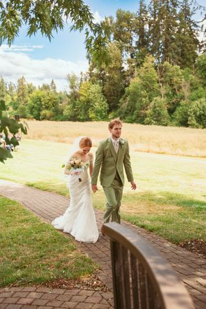 Couple shares a playful moment walking down a brick pathway surrounded by green trees on their wedding day.