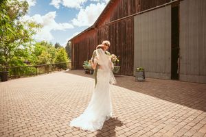 A figure in a flowing white wedding dress dances and spins gracefully on a brick patio near a rustic brown barn.