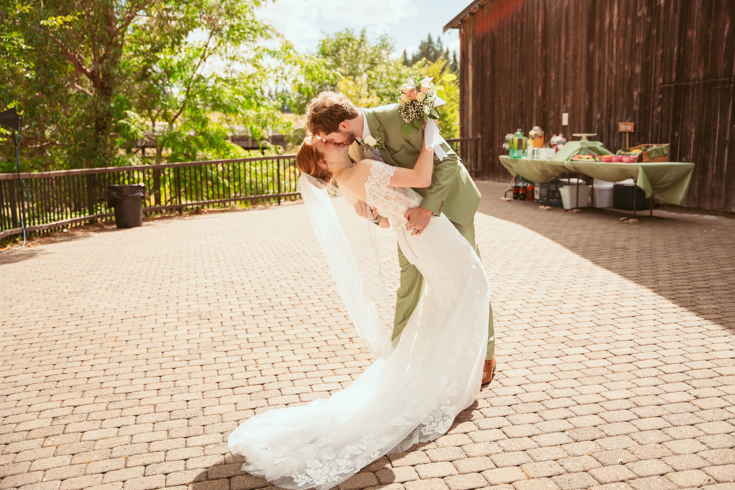 A couple in wedding attire dances and dips gracefully on an outdoor brick patio surrounded by lush greenery.