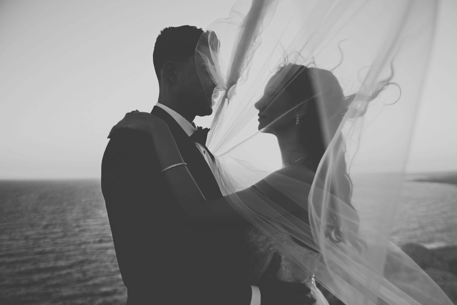 Dreamy black and white silhouette of a bride and groom standing together near a waterfront with flowing veil.