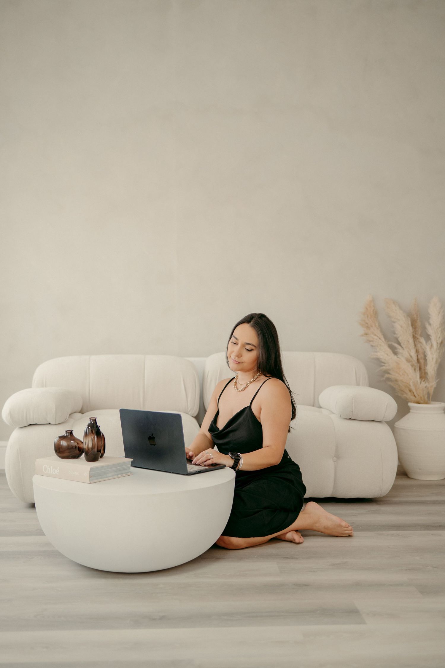 Woman in black lounging comfortably against a white curved chair while working on a laptop.