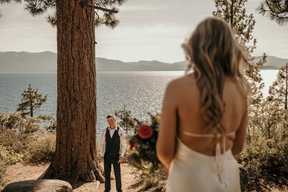 A romantic first look moment beneath towering pine trees overlooking a serene lake.