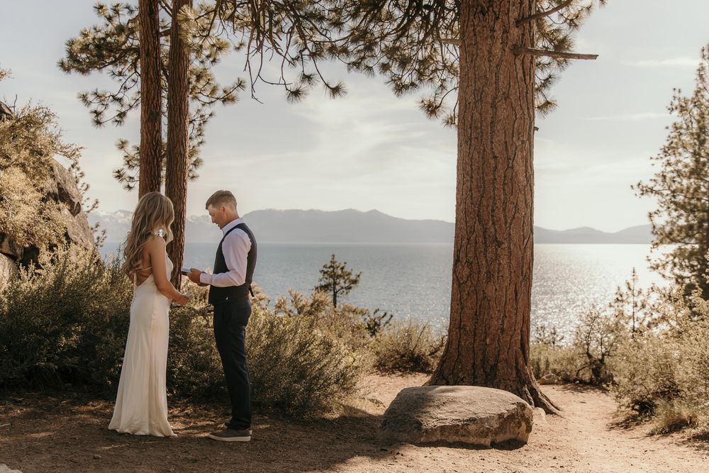 A couple shares an intimate moment by a lake surrounded by tall pine trees during a sunset ceremony.