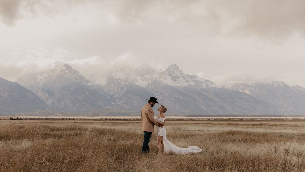 A couple stands in a field with the majestic Teton mountains creating a dramatic backdrop.