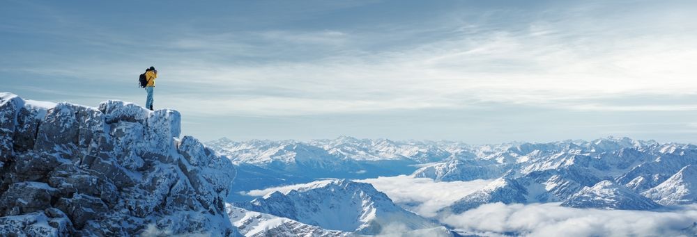 Lone figure standing on snowy mountain peak overlooking vast mountain range under cloudy sky.