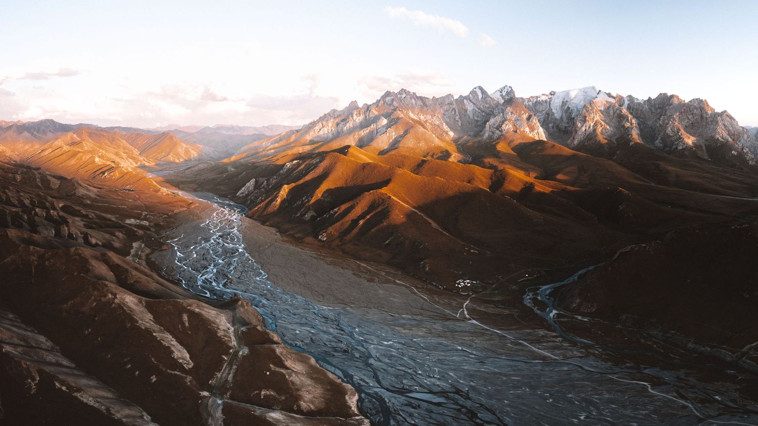 Snow-capped mountain peaks rise against a clear sky with sunlight casting golden hues on the rugged terrain.