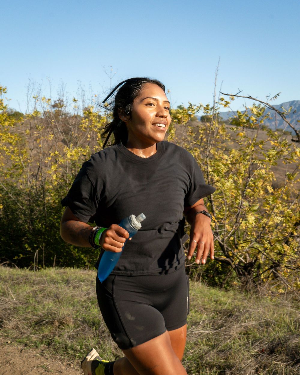 Runner in black outfit holding a blue soft flask while running on a sunny hillside trail.