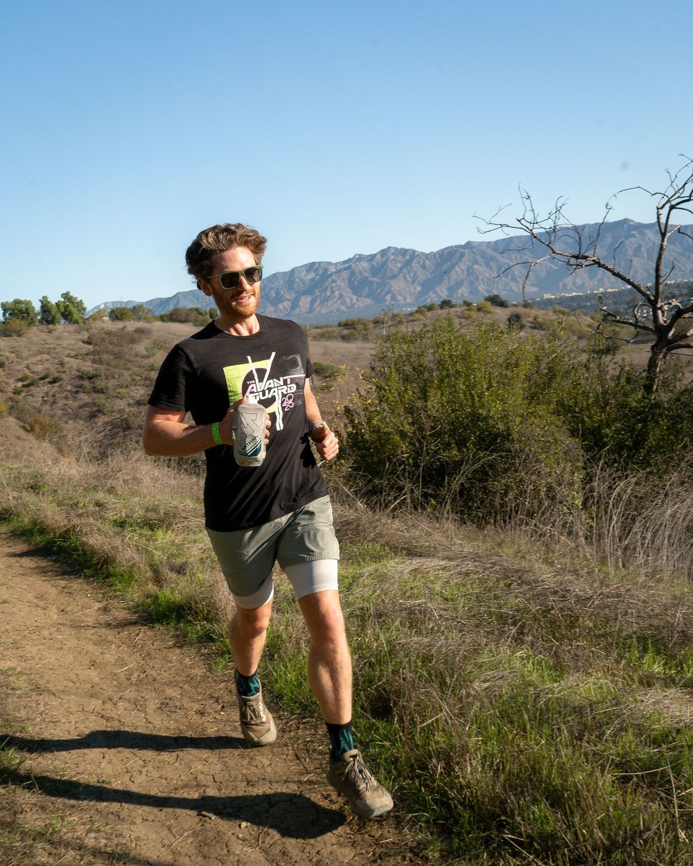 Runner in black T-shirt and gray shorts jogging on a hillside dirt trail with mountains behind.