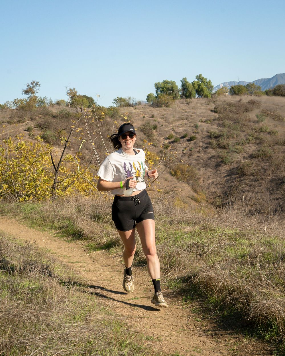 Runner in white T-shirt and black shorts jogging along a winding dirt trail in dry hills.