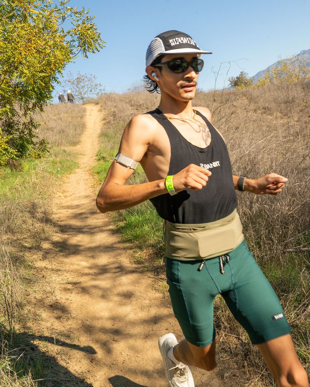Runner in tank top and shorts on a dirt trail through dry grass under a clear sky.