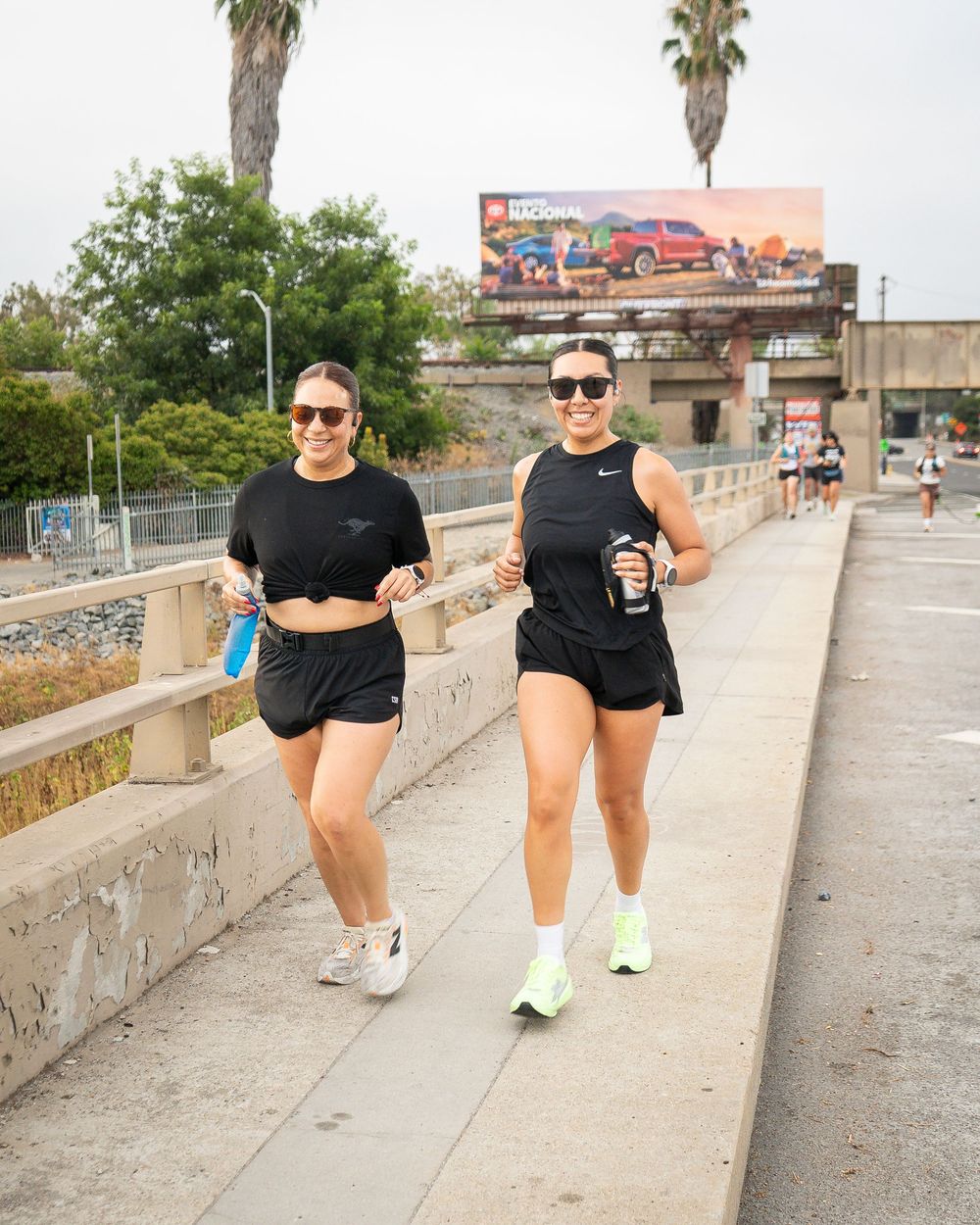 Two runners in black tops jog side by side on a bridge holding water bottles.