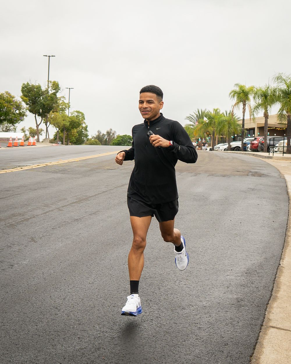 Runner in black outfit jogs alone on a wet street lined with palm trees and parked cars.