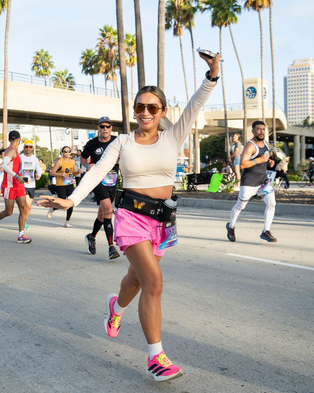 Marathon runner in pink shorts raises arms joyfully while running with a crowd on a palm-lined city street.