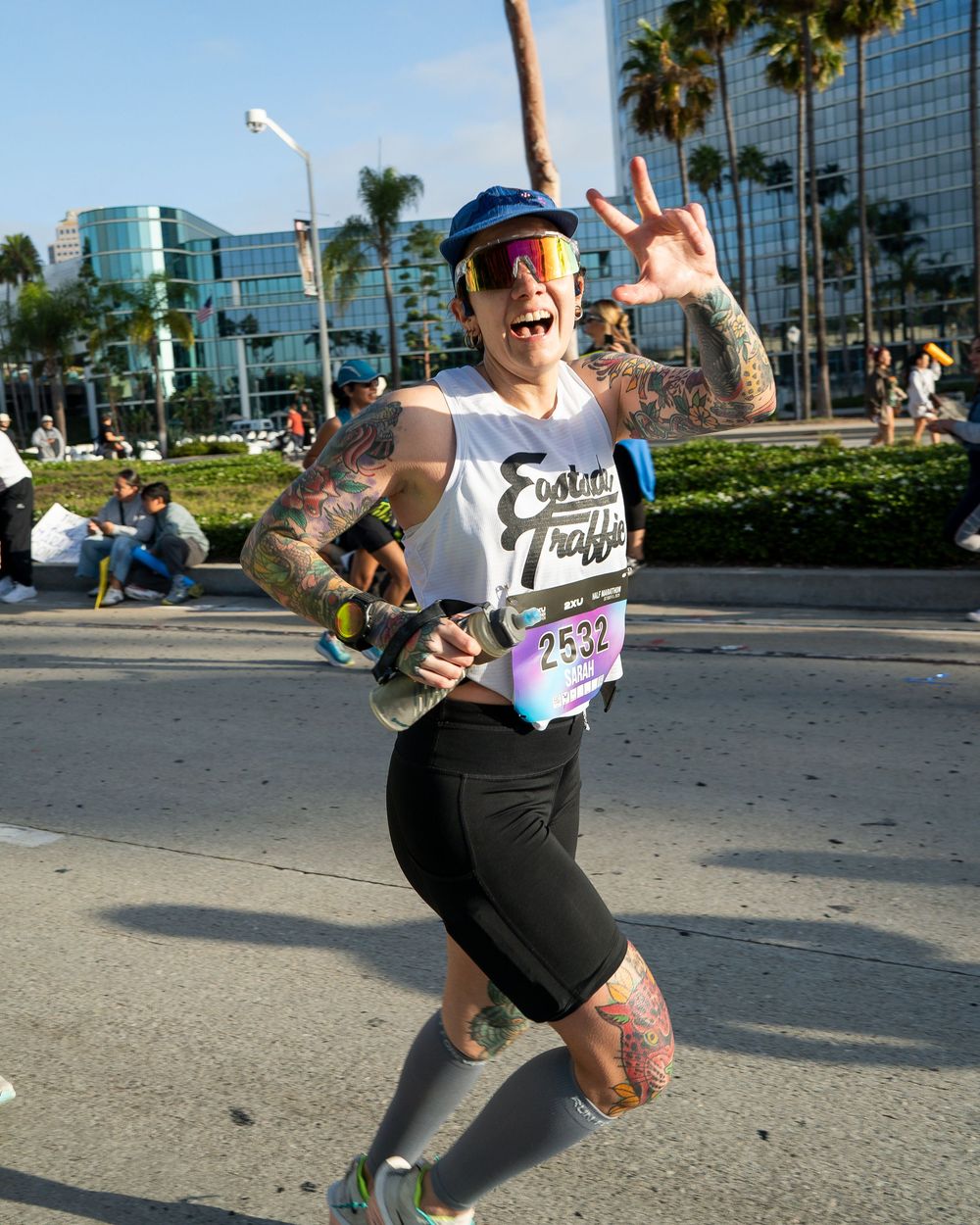 Tattooed marathon runner in race bib smiles and flashes a hand sign while running on a city street.