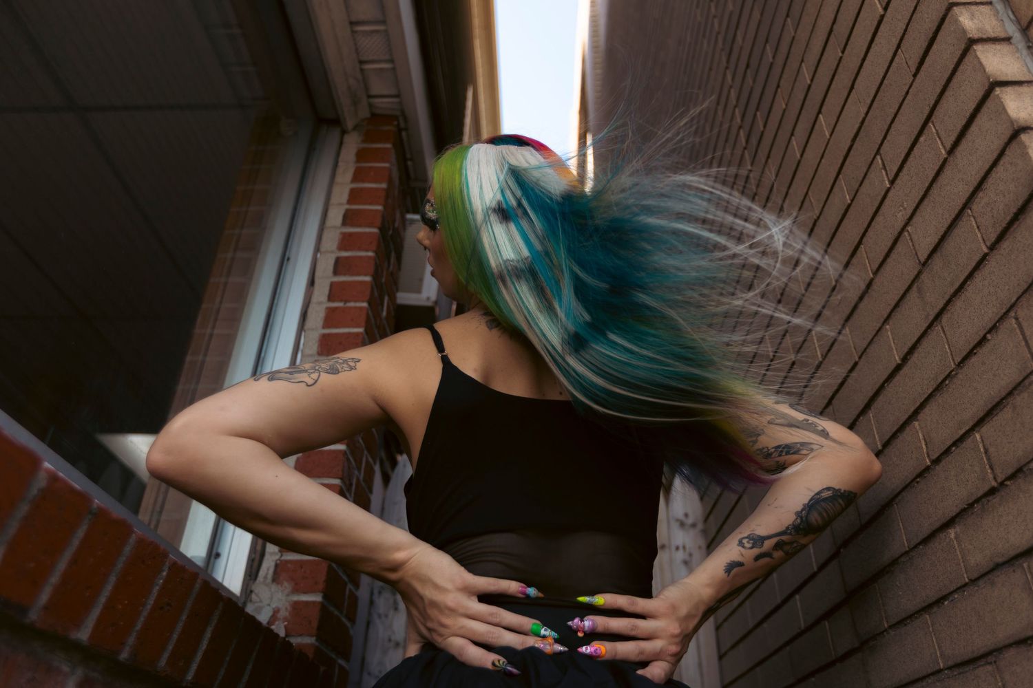 Person with vibrant turquoise hair wearing black clothing stands against a brick wall in an outdoor corridor.