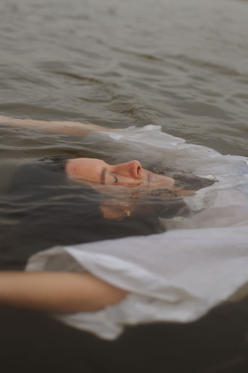 A person floats serenely in dark water wearing white fabric that creates an ethereal underwater effect.