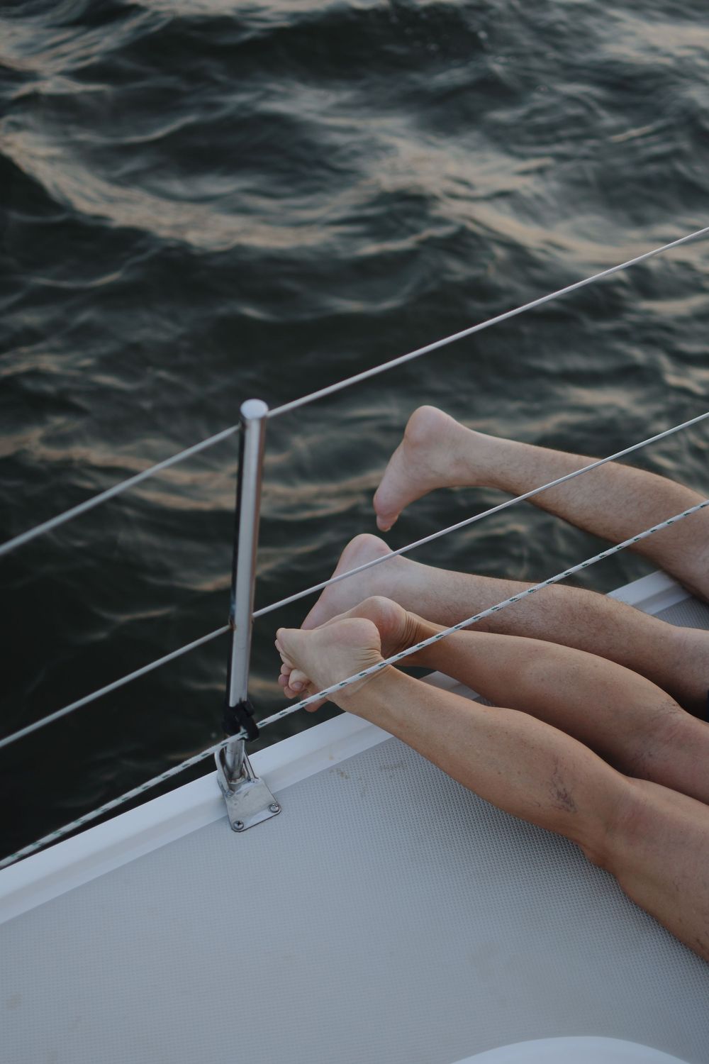 Bare feet relaxing on the edge of a sailboat with metal safety cables visible against dark ocean waters.