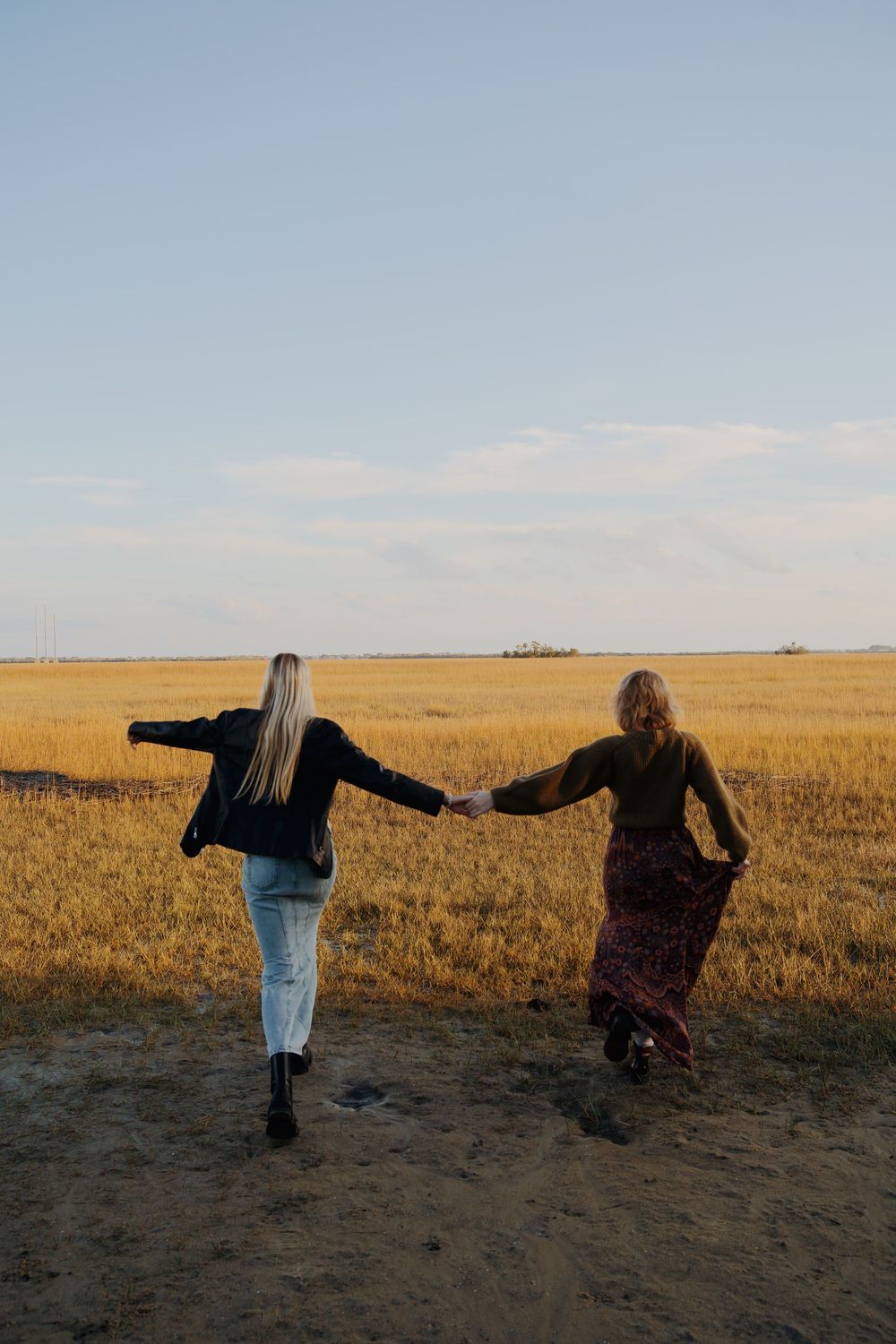 Two friends hold hands while walking through a golden wheat field at sunset, creating a carefree silhouette against the horizon.
