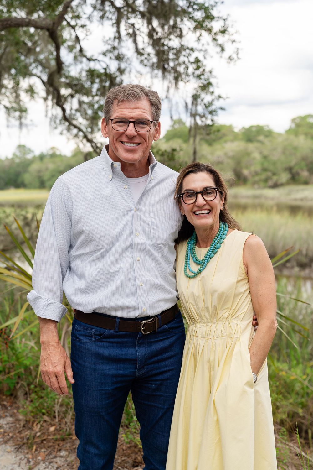 A couple stands together outdoors, wearing glasses and casual attire against a natural landscape with trees and water.