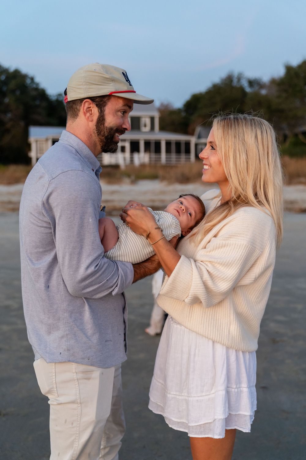 A couple in white and gray outfits share playful candid moments during a beach photoshoot at sunset.