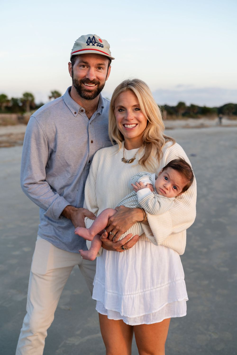 A family in coordinated white and beige outfits poses together for casual beach photos at sunset.
