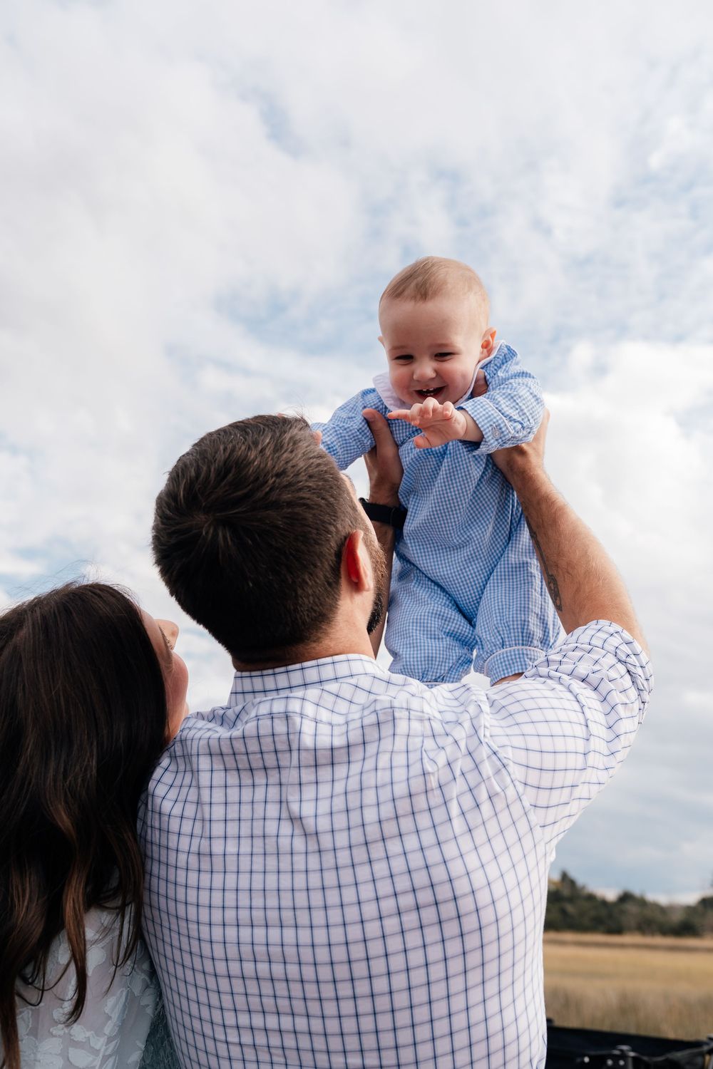 A parent lifts a joyful baby wearing light blue against a cloudy sky background in an outdoor setting.