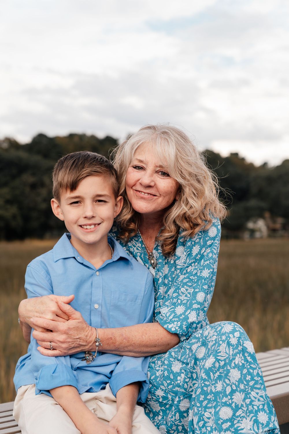 A woman in a blue floral dress embraces a young boy wearing a light blue shirt while sitting outdoors.