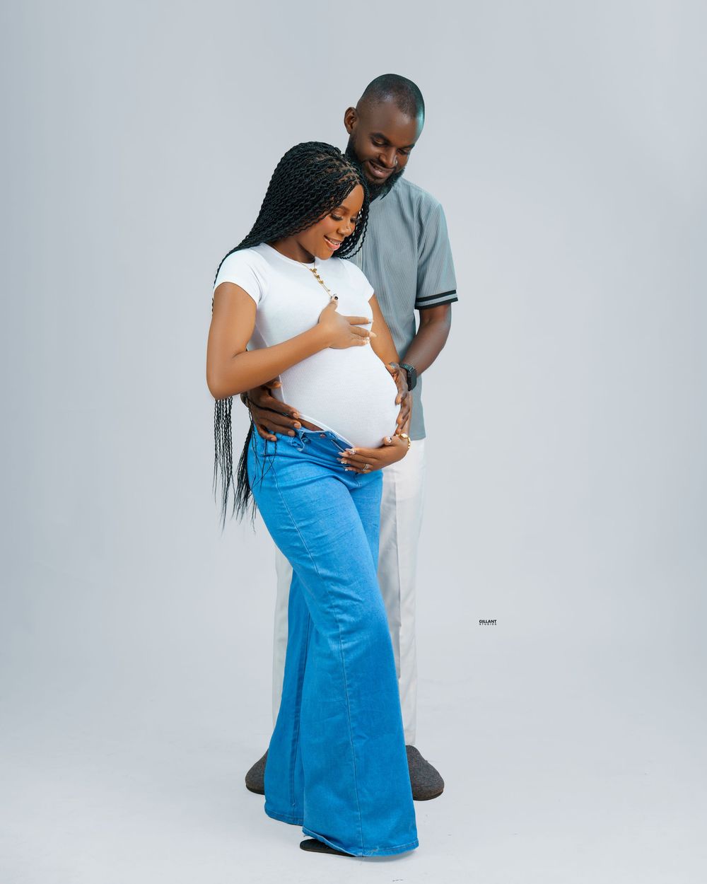 Tender maternity photo of expecting couple in casual wear against white backdrop.
