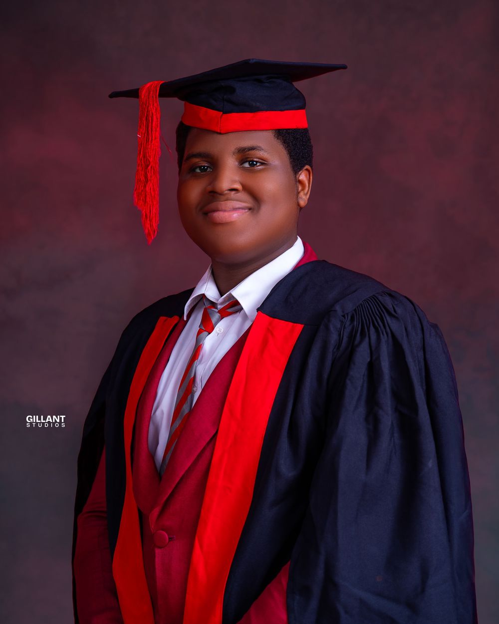 Graduate wearing academic regalia with black cap, navy gown, and red sash in formal portrait.