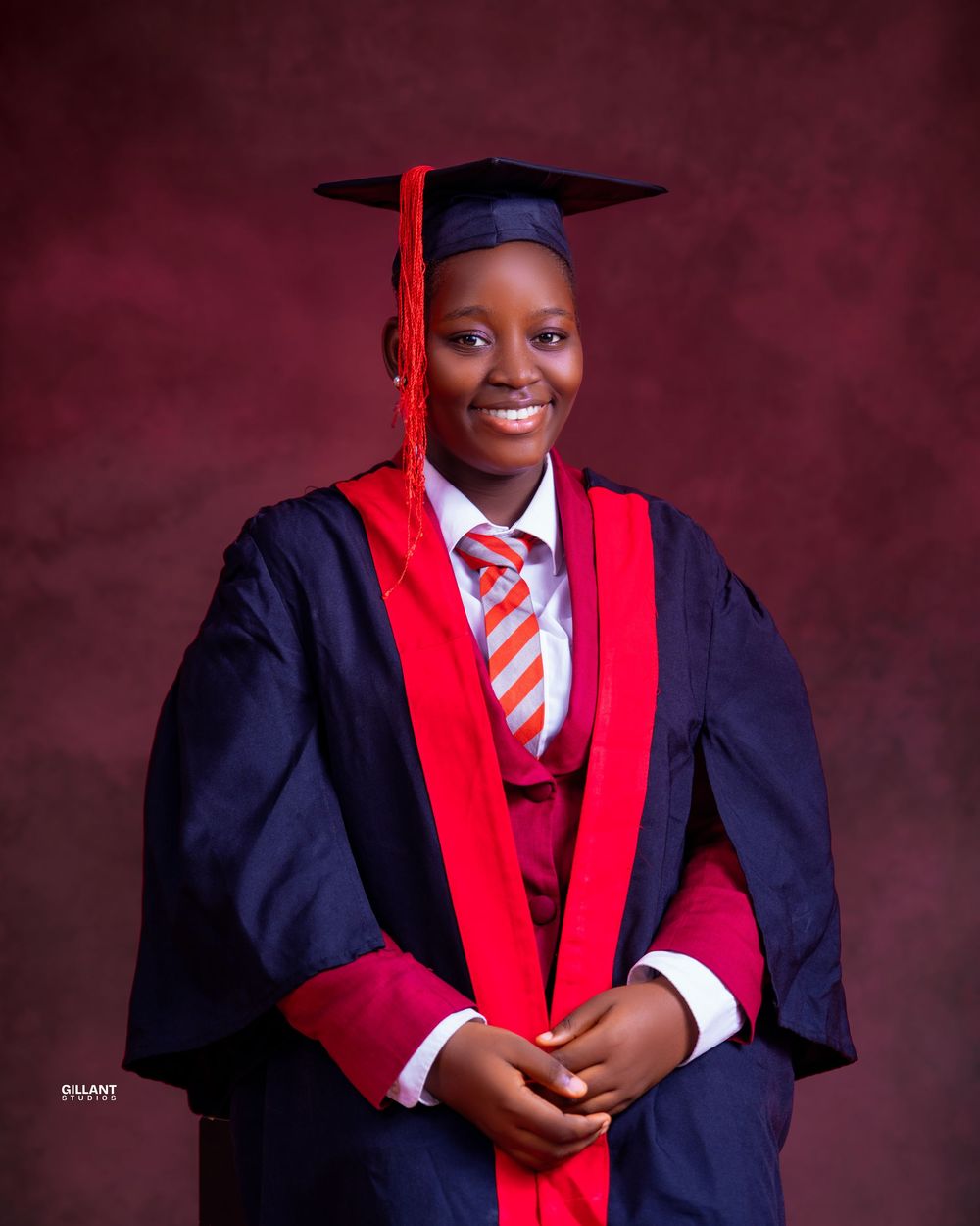Academic portrait in navy graduation gown with red accents and striped tie against burgundy backdrop.