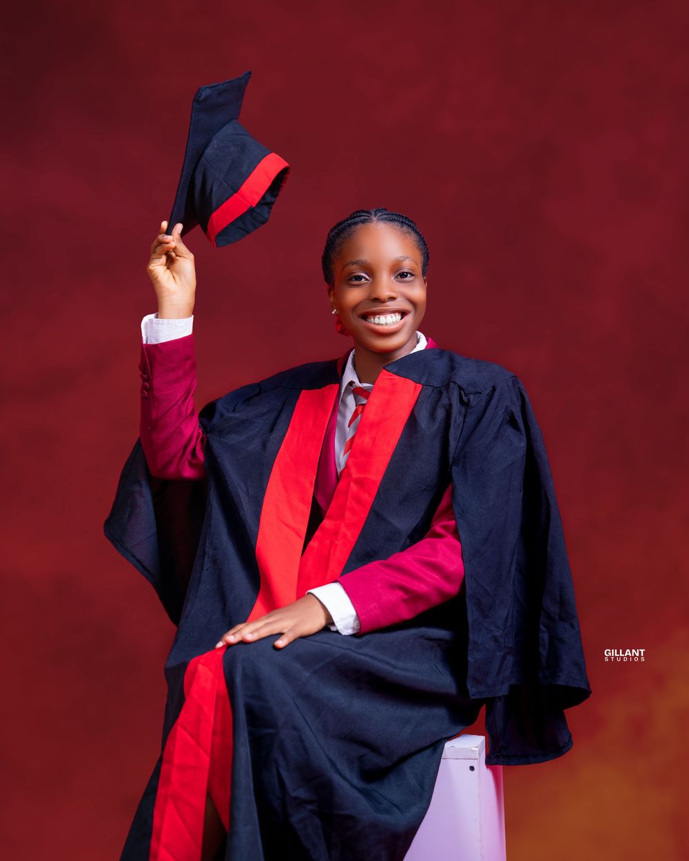 Graduate in navy and red academic regalia celebrates with raised cap against red background.