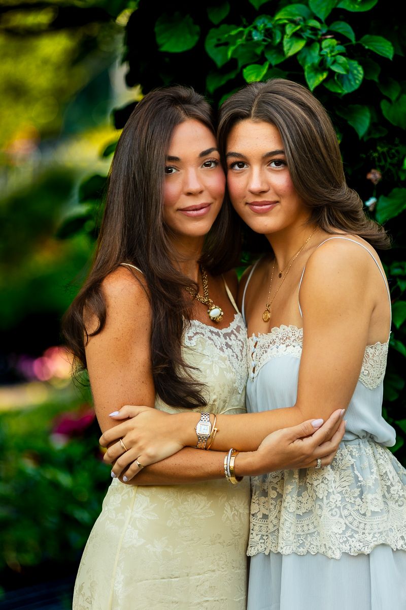 Two young women in elegant dresses pose together against a green garden background with flowers.