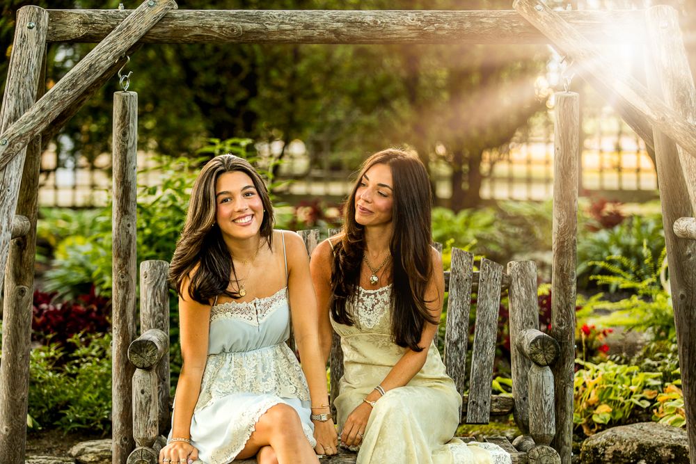 Two women in sundresses sit on rustic wooden bench in garden during golden hour sunset.