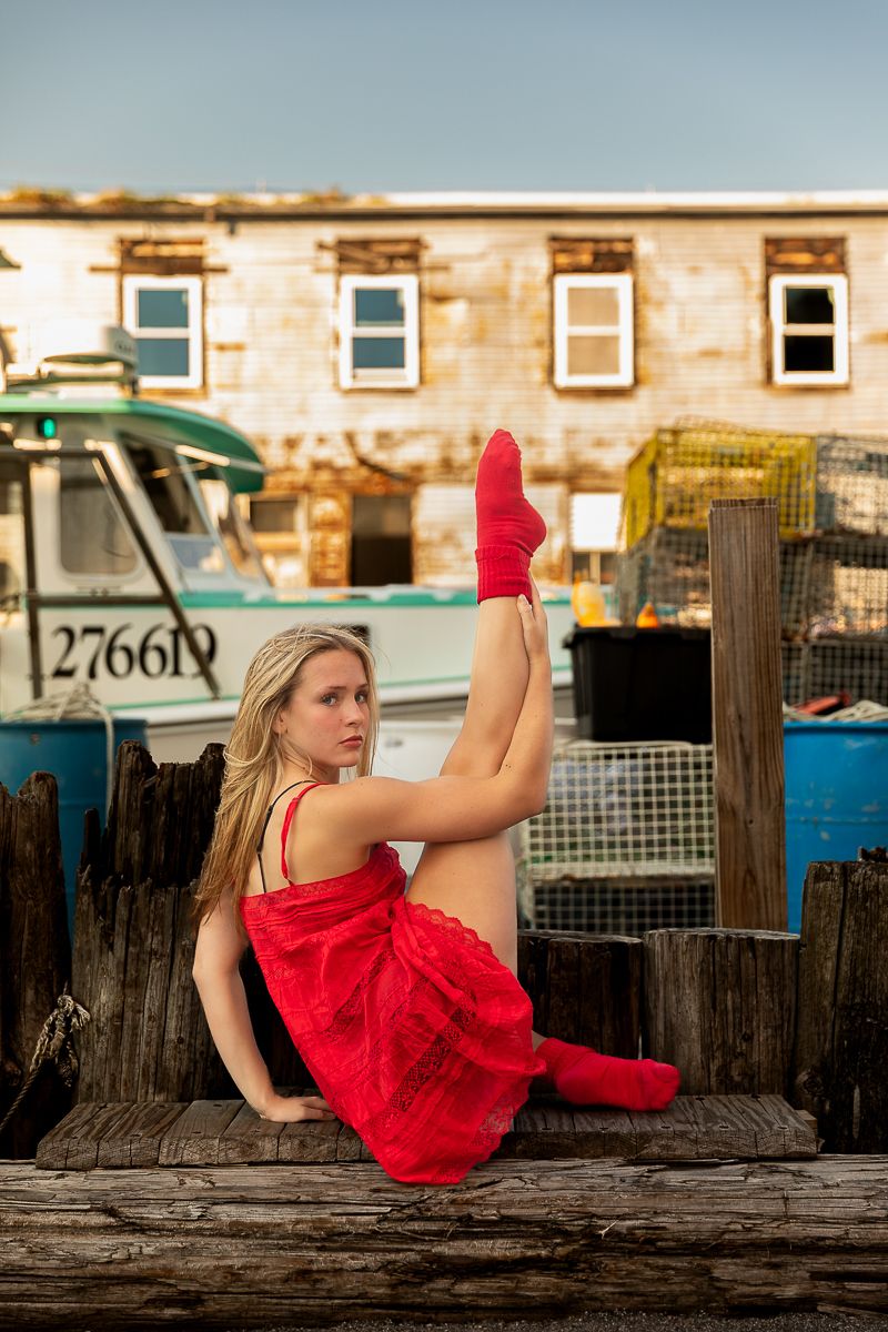 Person in a bright red dress poses on a wooden dock at sunset with boats and buildings in the background.