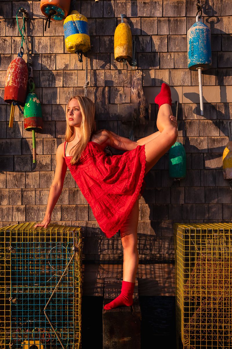 A ballet dancer in a red lace dress poses dynamically against a rustic wall decorated with colorful buoys.