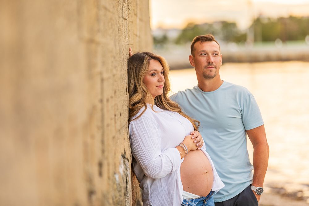Expecting couple poses against stone wall at sunset in casual attire during maternity photoshoot.