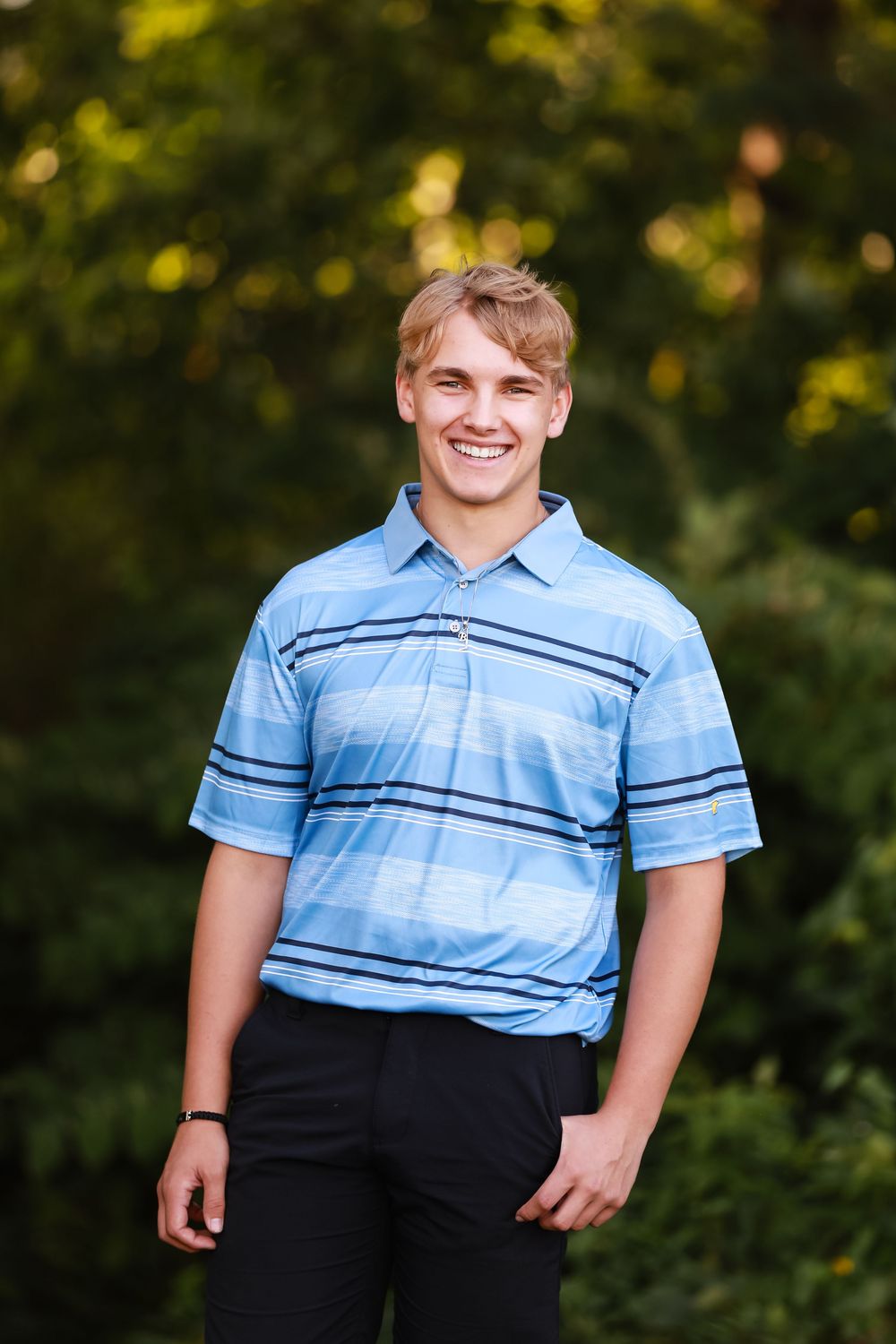 A person wearing a light blue striped polo shirt and dark pants poses against a blurred outdoor background with trees.