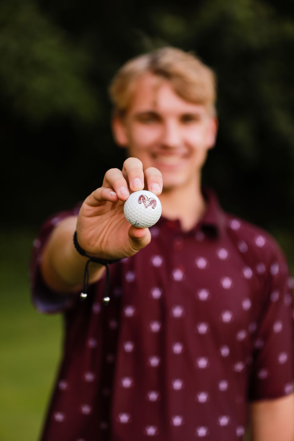 A person in a burgundy polka dot shirt holds up a golf ball in a grassy outdoor setting.