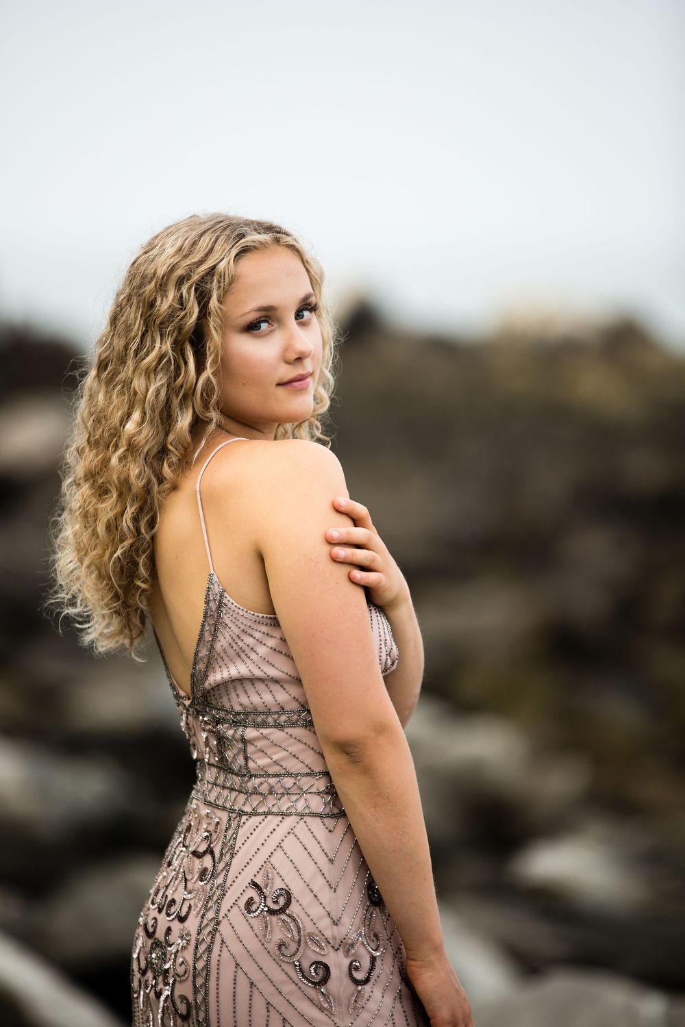 Elegant portrait poses in beaded dress against misty coastal background.