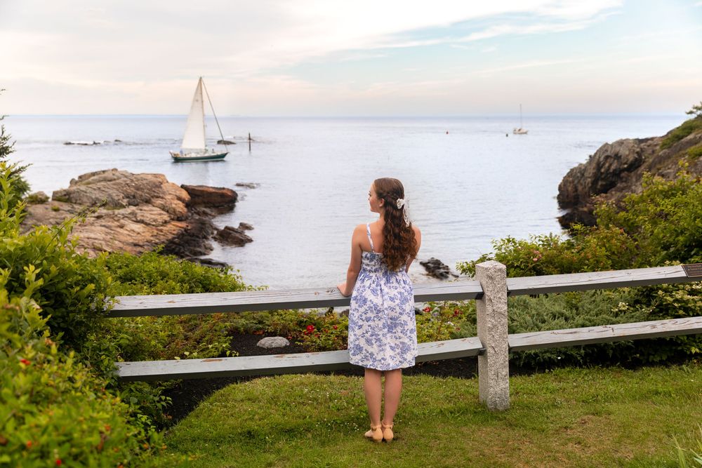 Someone in a floral dress watches sailboats from a coastal overlook on a serene evening by the sea.