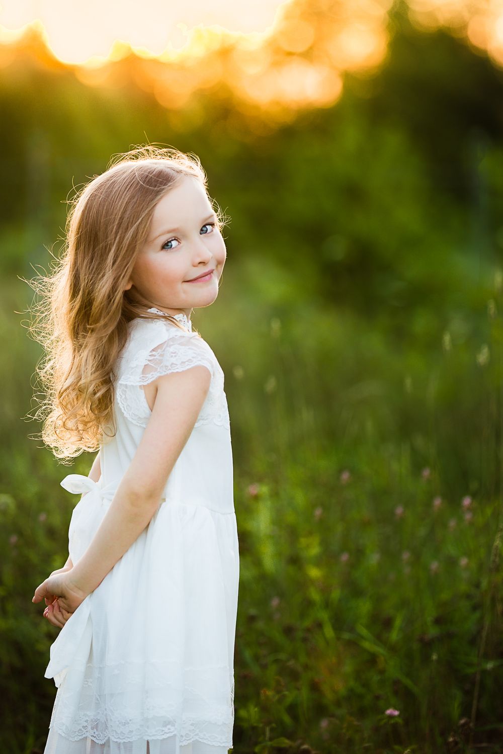Child in white lace dress smiles while twirling in a sunlit meadow during golden hour.