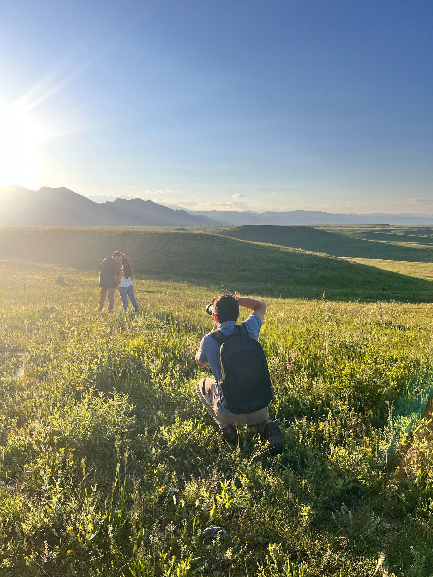 Photographer taking pictures at sunset on grassy hillside with mountain backdrop and golden sunlight.
