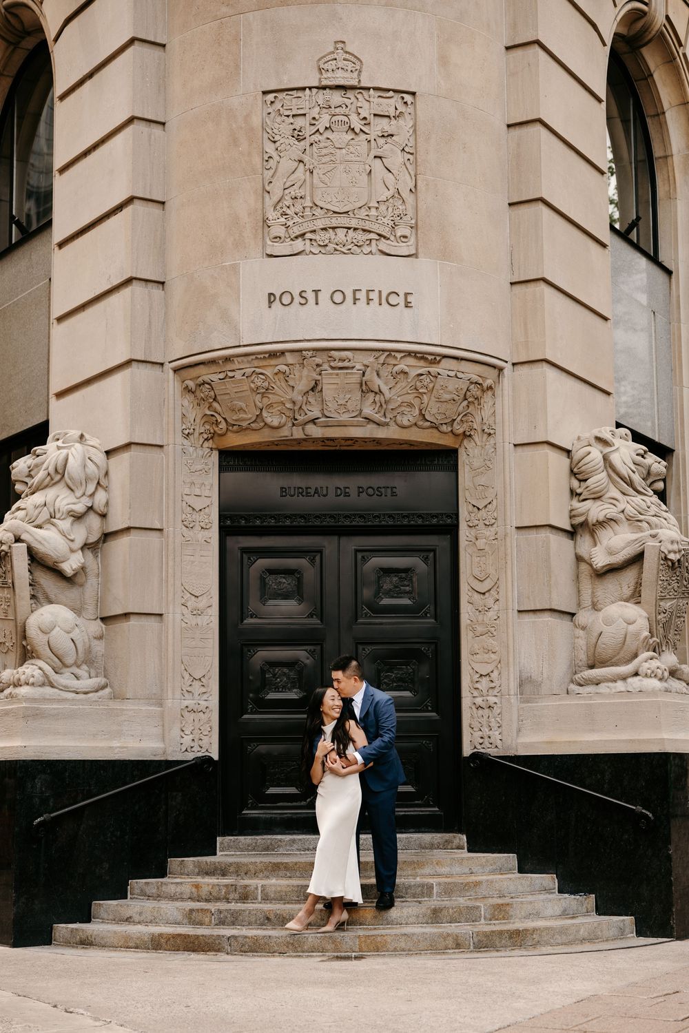 A couple shares a tender moment on the steps of a historic stone post office building with ornate architectural details.