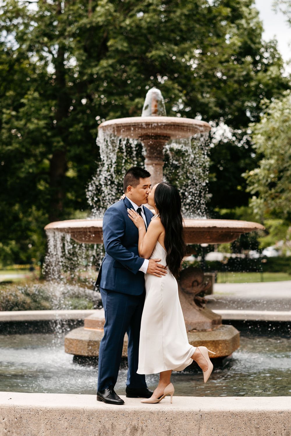 A romantic couple shares an intimate moment by a flowing fountain in a garden setting.
