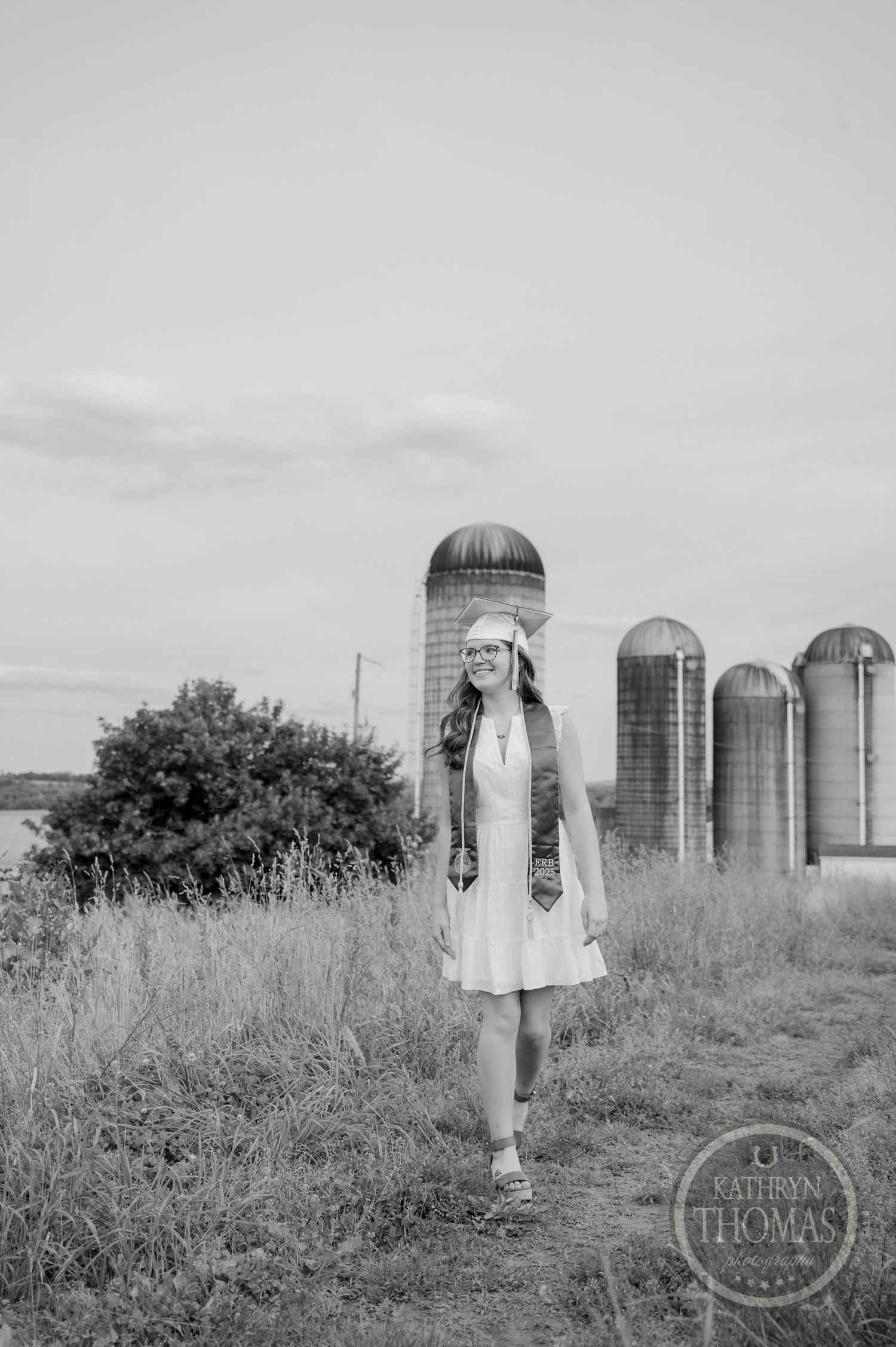 Black and white photo of a person in a dress standing in front of tall farm silos on a rural path.