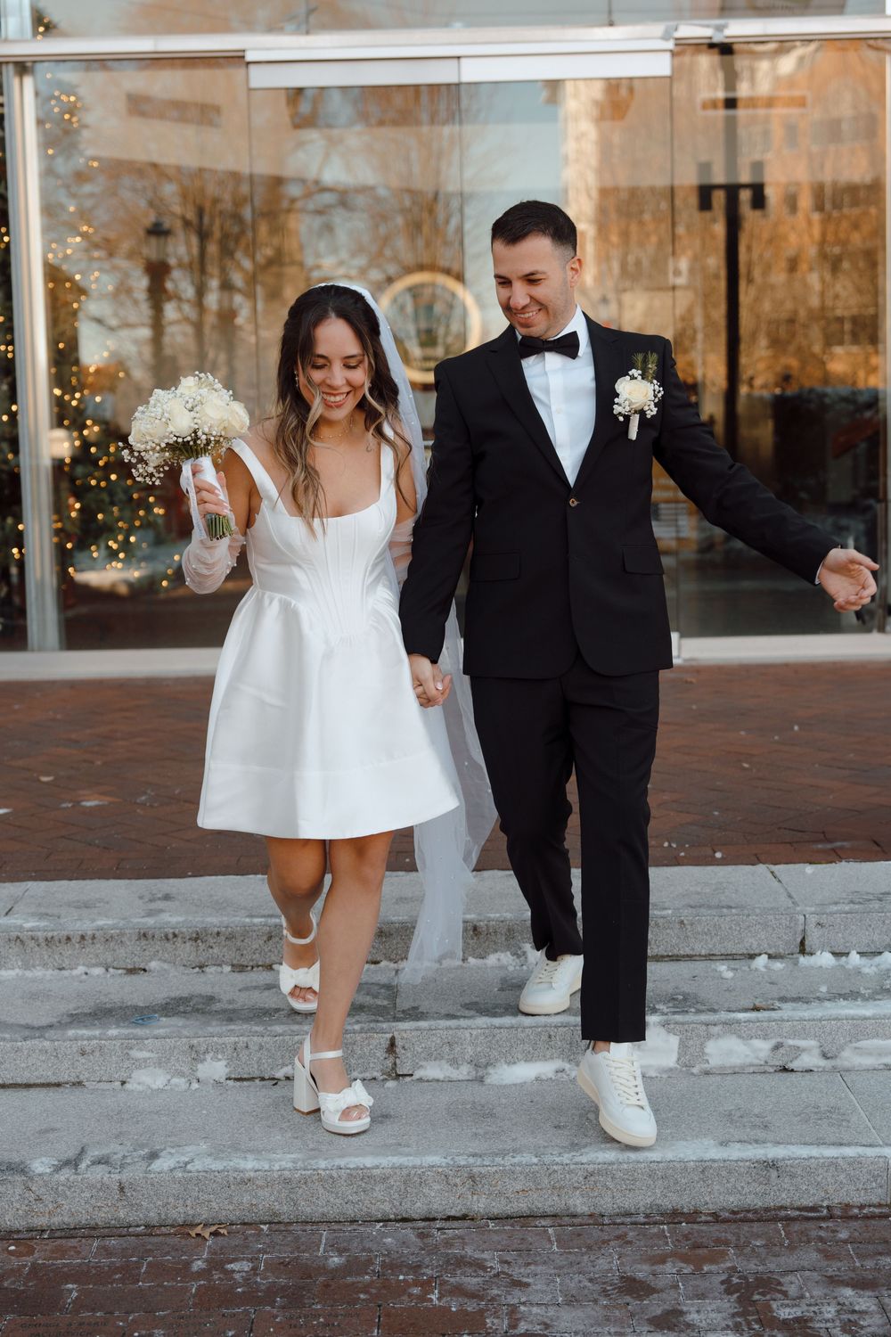 A couple descends brick stairs in matching white shoes, the bride in a short dress with a bouquet.